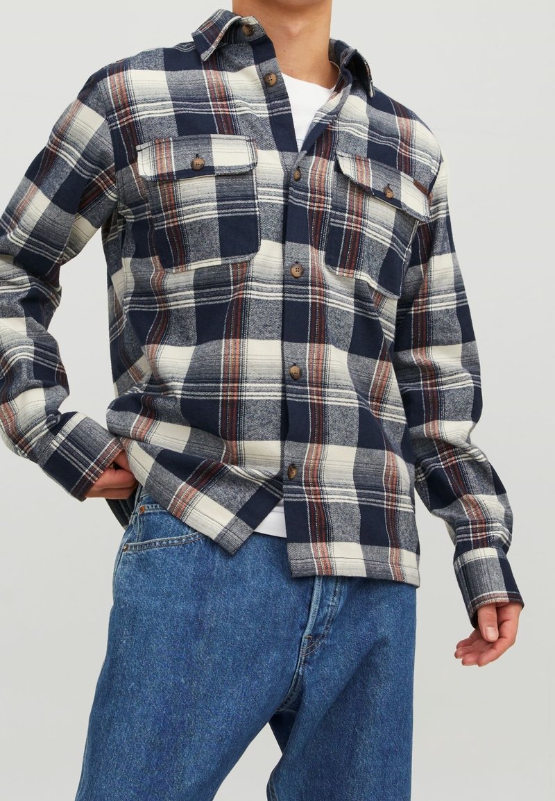 Camicia a quadri blu e crema con bottoni marroni, due tasche sul petto e colletto classico. Indossata con jeans di denim blu.