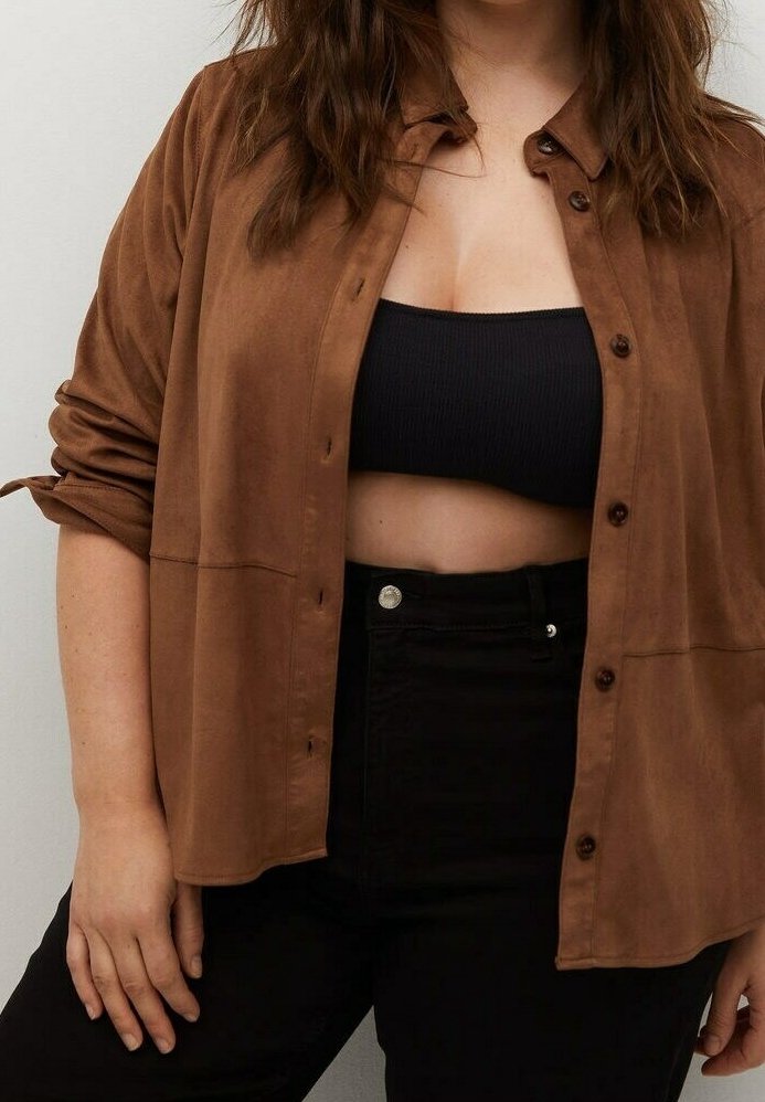 Femme portant une chemise marron boutonnée par-dessus un crop top noir et un pantalon noir taille haute, manches retroussées et cheveux lâchés.