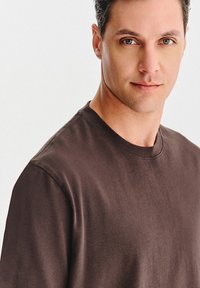 Mężczyzna z krótkimi ciemnymi włosami i jasnymi oczami, ubrany w prostą, ciemno-brązową koszulkę typu crew-neck, patrzy na kamerę z neutralnym wyrazem twarzy.