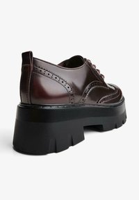 Scarpa brogue in pelle marrone scuro con suola piatta nera spessa e dettagliate perforazioni, mostrata dal lato posteriore.