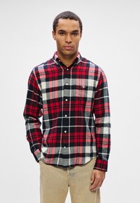 Hombre vistiendo una camisa de cuadros roja, negra y blanca con pantalones beiges, de pie contra un fondo claro, mirando de frente con las manos en los bolsillos.