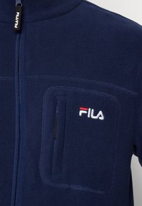 Giacca in pile di colore blu navy con zip frontale, taschino sul petto e dettagli logo in contrasto bianco e rosso. Texture morbida e design resistente.