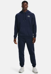 Dunkelblauer Hoodie mit einer Kängurutasche, Kapuze mit Kordelzug und weißem Logo. Kombiniert mit passenden dunkelblauen Jogginghosen und hellgrauen Sportschuhen.