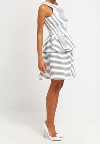 Bílo-náměsíčně pruhované bezramínkové šaty s peplum pasem a rozšířenou sukní, doplněné bílými vysokými podpatky.