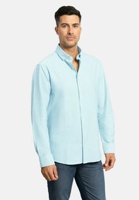 Camisa azul claro de botones, con mangas largas y un pequeño cuello, hecha de una tela suave con una textura sutil. Llevada con jeans oscuros.