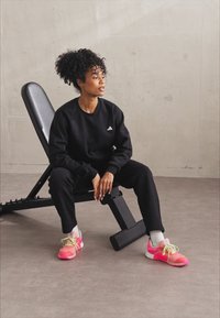 Μαύρο φούτερ με λευκό λογότυπο, ταιριαστά μαύρα παντελόνια και πολύχρωμα ροζ αθλητικά παπούτσια με πράσισα κορδόνια. Ουδέτερο φόντο με έναν πάγκο γυμναστηρίου.