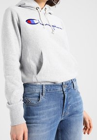 Sudadera de felpa gris con bolsillo frontal, capucha con cordón y logotipo bordado en azul y rojo. Combinada con jeans de mezclilla azul claro con costuras decorativas.