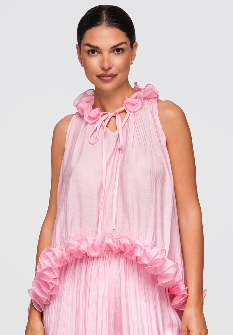 Femme aux cheveux foncés portant une robe rose clair sans manches avec col et ourlet à volants, nouée d'un nœud au niveau du cou, sur fond uni.