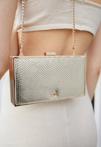 Metaliczna srebrna torebka kopertówka o fakturze węża, w prostokątnym kształcie, z złotym detalem sprzętowym, wyposażona w łańcuszek do noszenia.