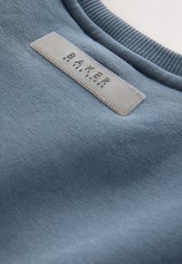 Blauer Sweatshirt-Stoff mit geripptem Kragen. Verfügt über ein rechteckiges Etikett, das mit "BAKER by TED BAKER" in Schwarz auf einer glatten, strukturierten Oberfläche geprägt ist.