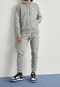 Sudadera gris de Nike con bolsillo frontal, combinada con pantalones de chándal grises. Zapatillas Nike en blanco y negro. Tejido suave, corte atlético, con un branding mínimo.