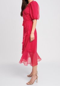 Eine Frau in einem roten Midikleid mit weißen Punkten, Puffärmeln, Rüschensaum und beigefarbenen, hochhackigen Sandalen mit Knöchelriemen, die seitlich steht.