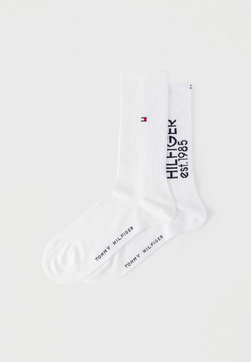 Δύο ζεύγη λευκών κάλτσων πλήρους μήκους με ριμπ μανσέτες, το ένα με ένα μικρό κόκκινο, λευκό και μπλε λογότυπο, το άλλο με το κείμενο "HILFIGER est. 1985".