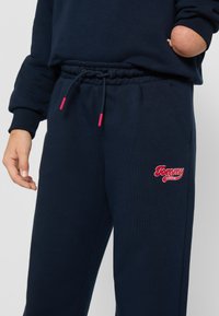 Jūras spēku sporta bikses ar gumiju jostasvietā, sarkanu auklu un logo uz kreisā augšstilba. Mīksta, teksturēta auduma bikses ar sānu kabatām.