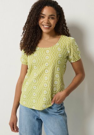 Jonge vrouw met krullend haar, een groen blouse met patroon en korte mouwen, en lichtblauwe spijkerbroek, glimlachend met één hand in de zak.