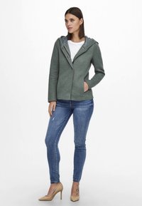 Green wool-blend jacket with a gray hood, featuring a collar, buttons, and front pockets, paired with blue skinny jeans and beige heels.