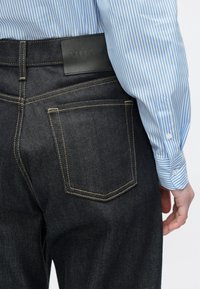 Jeans din denim închis la culoare, cu cusături galbene și o etichetă din piele neagră în talie, asortate cu un mânecă de cămașă în dungi albastre și albe.