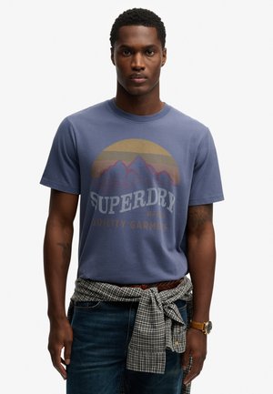 Mężczyzna w niebieskiej koszulce Superdry z grafiką gór, w kratę koszula przewiązana w talii, ciemne jeansy i złoty zegarek na rękę na białym tle.