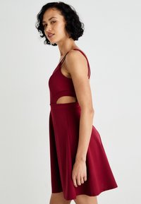 Burgundowa sukienka bez rękawów z dopasowanym gorsetem, wycięciami w talii i rozkloszowanym dołem. Wykonana z gładkiego, lekkiego materiału.