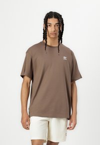 Brūns kokvilnas T-krekls ar brīvu piegulšanu, īsām piedurknēm un baltu Adidas logo kreisajā krūtīs. Saskaņots ar gaiši krēmkrāsas šortiem.