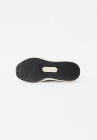 Sort gummisål med struktureret slidbane, der har en lysegrå kant og et centralt logo præget på en beige accent.