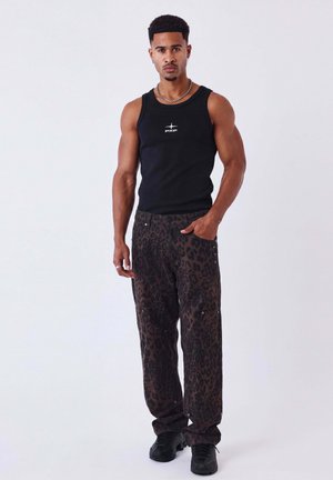 Giovane uomo con capelli corti e ricci che indossa una canotta nera e pantaloni scuri con stampa leopardata, in piedi su fondo bianco semplice.