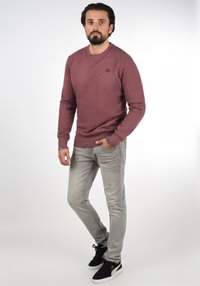 Pulover bordo cu mâneci lungi, gât rotund și logo mic, asortat cu blugi gri deschis și adidași negri cu accente aurii.