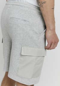 Shorts en coton gris avec une texture tissée, présentant une poche cargo latérale en gris clair et une poche arrière standard. Taille élastique.