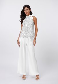 White lace top with halter neck and bow accent, featuring scalloped hem; paired with loose-fitting white trousers and strappy sandals.