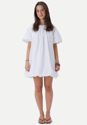 Jeune femme aux longs cheveux bruns portant une robe blanche ample à manches courtes et des sandales marron, debout devant un fond uni.