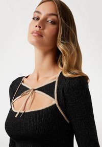 Top nero a costine con design a taglio, presenta una rifinitura beige e una chiusura a cravatta sul collo. Texture morbida con una leggera brillantezza.