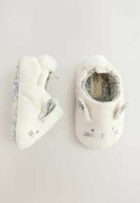 Pantuflas de bebé de algodón en color blanco roto con orejas de conejo y detalles faciales bordados. Forro con estampado floral y acentos de pom-pom. Textura suave, forma redondeada.