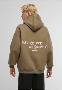 Brun oversized hettegenser med snøring, ribbestrikkede mansjetter og hvit grafisk tekst "KAFFEDATE PÅ SØNDAG?" på baksiden.
