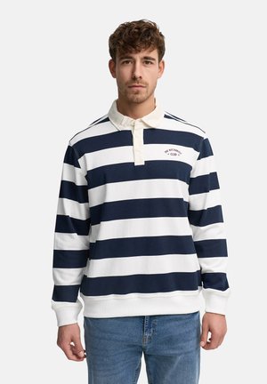 Młody mężczyzna z krótkimi kręconymi włosami, ubrany w granatowo-białą pasiastą koszulę rugby z długim rękawem oraz niebieskie dżinsy, stoi na tle gładkiego tła.