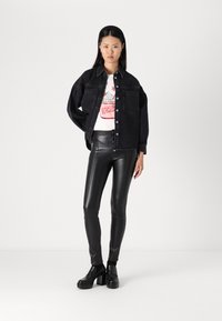 Musta denim-takki rento istuvuudella, valkoinen graafinen t-paita alla, jossa on kakku-kuvio, ja hyvin istuvat mustat nahkahousut kiiltävällä pinnalla.