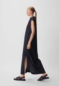 Langes, dunkelblaues ärmelloses Kleid mit einem Kragen, seitlichen Schlitzen und einer Brusttasche. Kombiniert mit dunklen Sandalen mit überkreuzten Riemen.