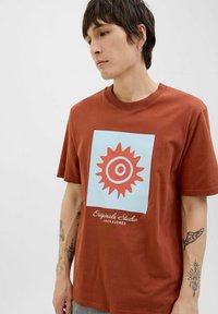 Brun bomulds t-shirt med et lysblåt grafisk design, der har en orange sol og teksten "Originals Studio JACK&JONES" nederst.