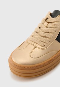Sneaker in pelle sintetica dorata con punta tonda, suola in gomma tan strutturata e lacci beige. Presenta un accento nero sul lato.