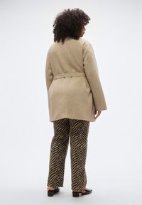 Smėlio spalvos vilnonis paltas su diržu liemens srityje, derinamas su juodomis ir rudu zebros dryžuoto audinio kelnėmis. Paltas turi ilgomis rankovėmis ir tiesų kirpimą.