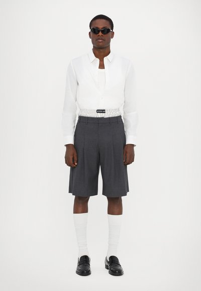 Biela košeľa s button-up zapínaním, šedé plisované šortky s elastickým pásom, biele ponožky a čierne mokasíny; slnečné okuliare dotvárajú celý outfit.