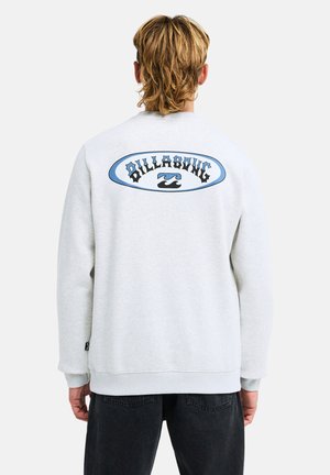 Osoba z blond włosami, ubrana w jasno szarą bluzę Billabong z niebieskim logo i czarne spodnie, widziana od tyłu.