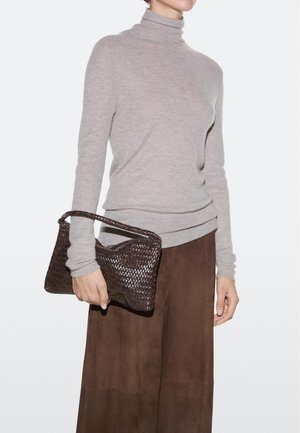 Persona con dolcevita beige e gonna marrone che tiene una borsa clutch intrecciata marrone scuro all'altezza della vita.