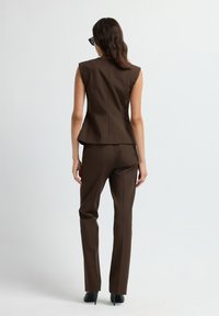 Vrouw in een donkerbruine mouwloze blazer en bijpassende broek, staand met de rug naar de camera tegen een effen lichte achtergrond.