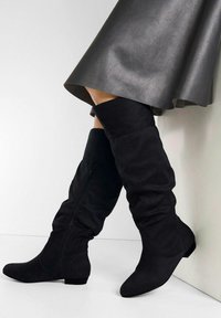 Bottes noires en suédine haute, à la tige fléchie, dotées d'un petit talon carré et d'une fermeture éclair sur le côté.