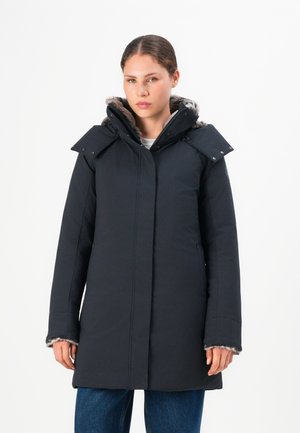 Vrouw in een lange zwarte winterjas met een bontgevoerde kap, staand tegen een witte achtergrond, met ontspannen handen langs haar sides.