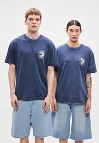 Tamsiai mėlynos medvilninės marškinėliai su apvalia iškirpte ir trumpomis rankovėmis, kairėje krūtinės pusėje su apvaliu logotipu. Derinami su šviesiai mėlynais džinsiniais šortais.