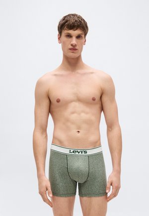 Giovane uomo con capelli corti castani che indossa boxer Levi's grigi con una vita bianca, in piedi davanti a uno sfondo chiaro e semplice.