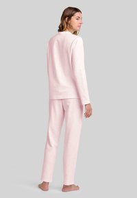 Mujer de pie descalza, de espaldas, con una blusa de manga larga rosa claro y pantalones a juego, frente a un fondo gris liso.