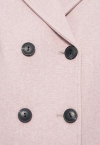 Δίπλα μπουφάν σε απαλό ροζ ύφασμα, με τέσσερα μαύρα κουμπιά, γιακάδες με εγκοπές και υφή που θυμίζει μάλλινη ύφανση.