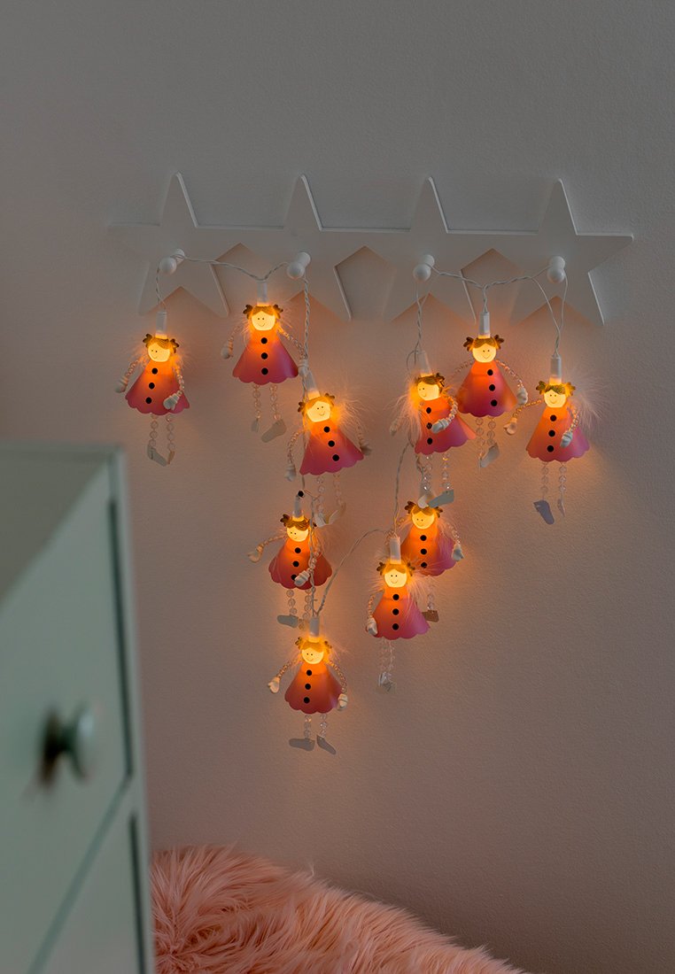 Lichterketten mit rosa, puppenförmigen Glühbirnen, von denen jede ein lächelndes Gesicht und Zöpfe hat, hängen von einem weißen, sternförmigen Halter an einer Wand.